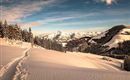 A snowy landscape with snow-covered trees and gentle hills. The sky is blue with some clouds, creating a tranquil winter atmosphere.