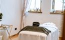 A quiet wellness room with a treatment table and large windows. The wooden floor and soft colors create a relaxed atmosphere.
