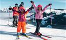 A group of skiers is standing on a snow-covered slope. Two excited children in colorful ski outfits are raising their hands in the air.
