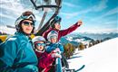 A family in a ski lift enjoying the view of the snow-covered mountains. They are wearing ski helmets and goggles and appear cheerful.