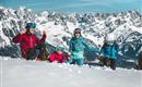 A family is playing in the snow with snow-covered mountains in the background. Everyone is wearing ski helmets and colorful winter clothing.