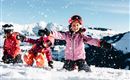 Three children are enthusiastically playing in the snow. They are laughing, throwing snowballs into the air, and wearing colorful ski clothing.