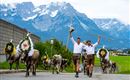 Two men in traditional attire lead a group of decorated cows through a picturesque landscape. Impressive mountains can be seen in the background.