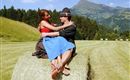 A couple is sitting on a hay bale in a beautiful, green landscape. The surroundings are hilly, with trees and a clear sky in the background.