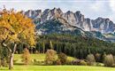 A picturesque landscape with majestic mountains and colorful autumn trees. In the foreground, there is a green meadow and a small house.