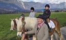 Two children are riding on ponies, accompanied by an older man. In the background, there is a green meadow and mountains under a blue sky.