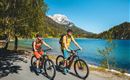 Two people are riding bicycles along a clear lake. In the background, there are mountains and blooming nature.