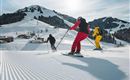 Three skiers are descending a groomed slope. In the background, there are snow-covered mountains and a ski lift visible.