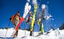Three skiers are standing in the snow and showing their skis. The sky is clear and blue, and fresh snow is being stirred up.