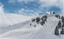 A snow-covered mountain landscape with skiers and pine trees. The sky is mostly blue with some clouds.