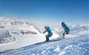 Two skiers are skiing down the snowy slope. In the background, there are snow-covered mountains and a blue sky.