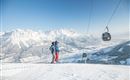 Ein Paar steht auf einem schneebedeckten Berggipfel mit Blick auf die winterliche Landschaft. Im Hintergrund sind die Berge und eine Seilbahn zu sehen.