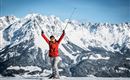 A person stands on a snow-covered mountain, lifting the ski poles into the air. In the background, impressive, snow-covered mountains can be seen.