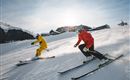 Two skiers are going down a snow-covered slope. One is wearing a yellow suit and the other a red one, while the sun shines in the background.