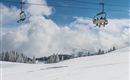 A snowy landscape with a ski lift and clouds in the background. The trees are covered in snow, and there is a clear, cold atmosphere.