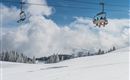 A snowy landscape with a ski lift and trees. The sky is clear and blue with some clouds.