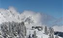 A snow-covered mountain landscape with tall fir trees and a ski lift. In the background, impressive mountains and blue sky can be seen.