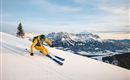 A skier in yellow gear is skiing on fresh snow. In the background, impressive mountains and a clear sky can be seen.
