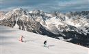 Zwei Skifahrer fahren eine verschneite Piste hinunter. Im Hintergrund sehen wir beeindruckende Berge unter einem bewölkten Himmel.