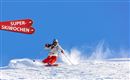 Ein Skifahrer fährt über frischen Schnee unter einem klaren blauen Himmel. Im Hintergrund sind Schilder mit der Aufschrift "SUPER-SKIWOCHEN" zu sehen.
