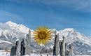 An impressive winter landscape with a sun sculpture and stone monuments. In the background, snow-covered mountains and a clear blue sky can be seen.