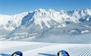 A snow-covered mountain landscape with a clear blue sky. In the foreground, two skis are lying on the groomed slope.