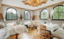 A cozy restaurant with wooden decor and large windows offering views of greenery. Tables are festively set and invite guests to linger.