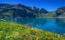 A clear lake surrounded by green mountains and colorful flowers. The sky is blue and sunny.