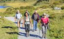A group of five people is hiking on a gravel path through a picturesque landscape. In the background, green hills and a blue lake can be seen.