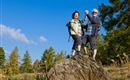 Two cheerful people are standing on a large stone outdoors. The sky is blue and there are trees in the background.