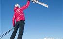 A person in a red ski jacket is lifting a pair of skis high. In the background, snow-covered mountains can be seen under a clear blue sky.