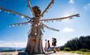 An impressive wooden sculpture in nature with radiant decorative elements. Two people and a dog are walking along a path in front of the scene.