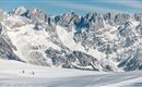 A snowy landscape with majestic mountains in the background. Two skiers glide over the glistening snow.