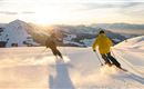 Zwei Skifahrer fahren bei Sonnenuntergang eine verschneite Piste hinunter. Im Hintergrund sind Berge und ein klarer Himmel zu sehen.