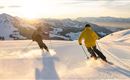 Two skiers are riding in the evening sun over a snow-covered slope. The mountains in the background provide a picturesque backdrop.
