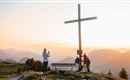 A group of people gathers around a large cross on a mountain. In the background, a picturesque mountain landscape can be seen at sunset.