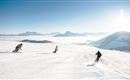 Three skiers enjoy a snow slope under a clear sky. Majestic mountains can be seen in the background.