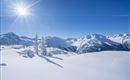 A snow-covered landscape with majestic mountains and a clear blue sky. The sun shines brightly over the winter scenery.