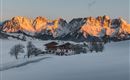 A picturesque winter landscape with snow-covered mountains and a traditional wooden house. The sun rays illuminate the mountain peaks in orange.