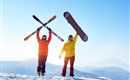 Two skiers are standing on a snow-covered mountain peak. They are holding their skis and a snowboard up in the air and enjoying the clear blue sky.