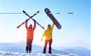 Zwei Wintersportler stehen auf einem schneebedeckten Berg. Einer hält Skier, der andere ein Snowboard hoch in die Luft.