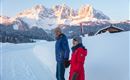 A couple in warm winter jackets stands on a snow-covered path. In the background, majestic mountains are visible, illuminated by sunlight.