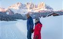Two people walk hand in hand on a snowy path. In the background, majestic mountains with snow-covered peaks can be seen.