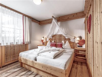 A cozy bedroom with a large wooden bed and white bedding. The room has bright windows and rustic wooden elements.