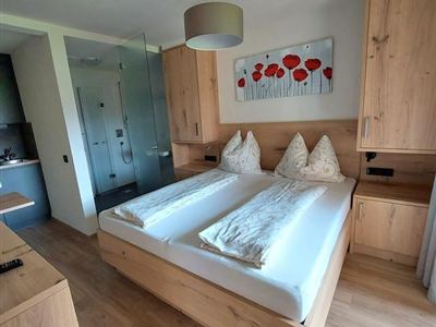 Ein modernes Schlafzimmer mit einem Doppelbett und weißen Bettdecken. Im Hintergrund befinden sich eine Dusche und eine Küchenzeile, sowie ein Bild mit roten Blumen an der Wand.