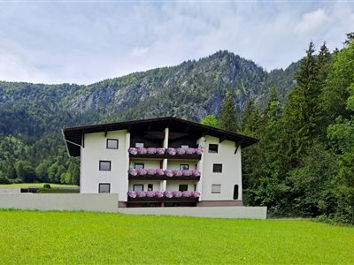 A modern building with multiple balconies stands in a green meadow. In the background, tall mountains and forests can be seen.