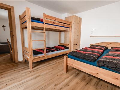 Een gezellige kamer met een stapelbed en een tweepersoonsbed. De vloer is van hout en de muren zijn licht geverfd.