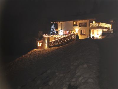A cozy house in the dark, festively lit with Christmas lights. In the foreground lies snow and a Christmas tree shines in the darkness.