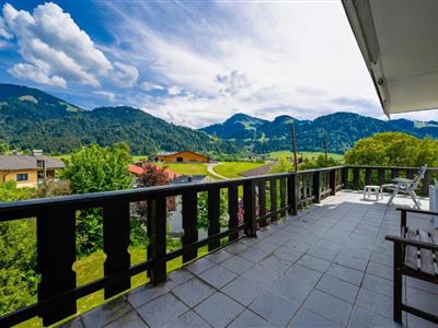 A terrace overlooking green mountains and a blue sky. In the foreground, there are chairs and a table.