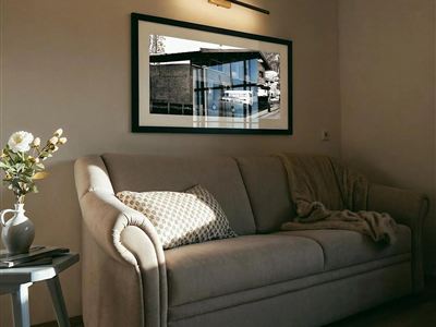 A cozy living room with a beige sofa and a warm light source. A framed picture hangs on the wall, depicting a modern building.